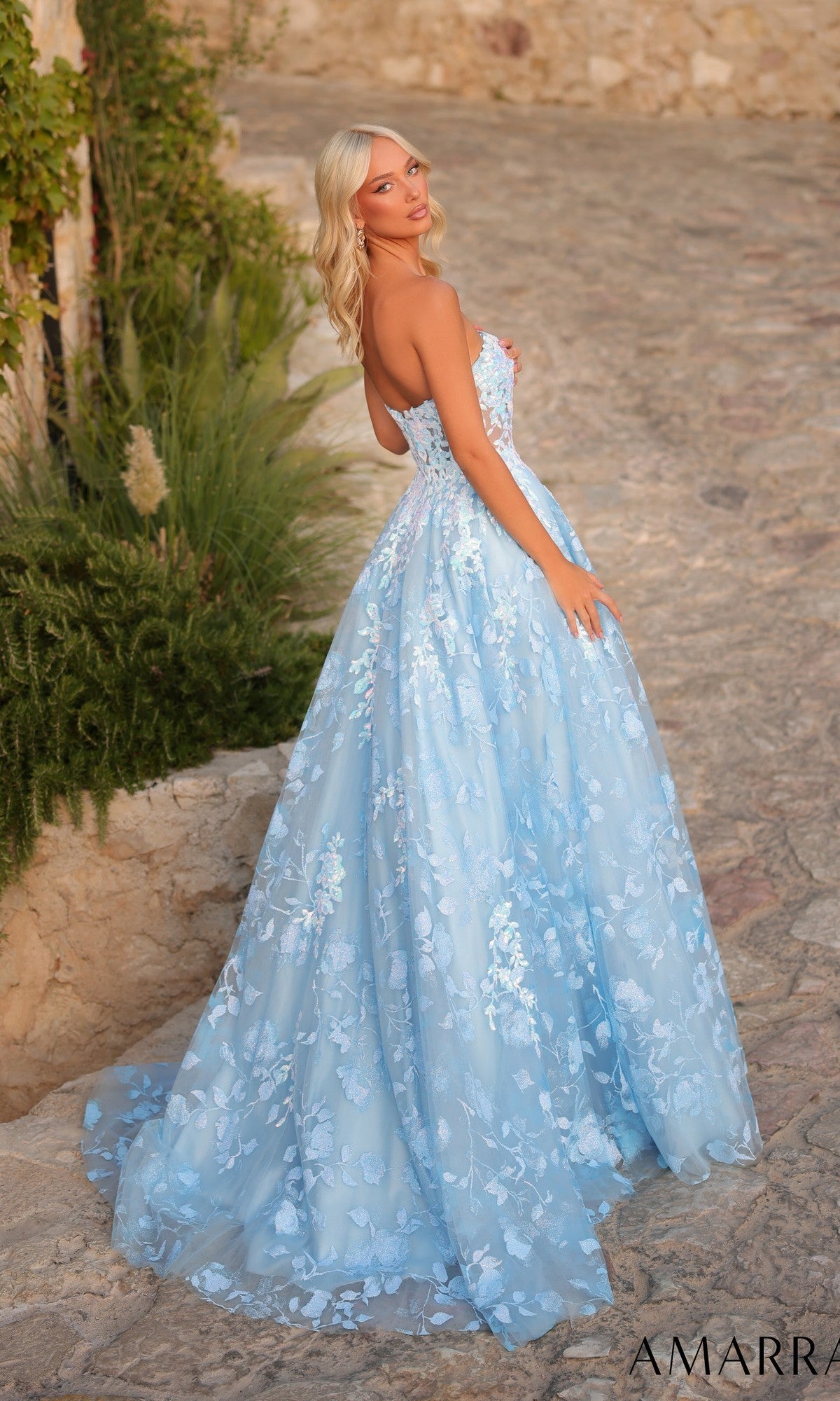 Wearing the Amarra 89513, a long light blue strapless prom dress with a floral-embroidered tulle skirt, a woman stands on a stone pathway and glances over her shoulder.